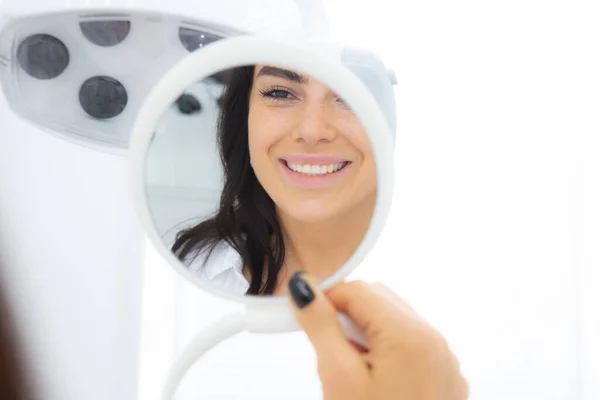 Mulher bonita olhando no espelho para o consultório odontológico desfrutando de seu belo sorriso e dentes após o tratamento odontológico na clínica odontológica, foco seletivo Imagem De Stock