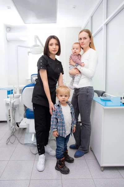 Bela jovem com um bebê em seus braços e um menino em uma clínica odontológica com um dentista pediátrico olhar para a câmera. primeira visita ao dentista. Visita de família ao médico — Fotografia de Stock