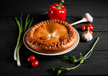 Round pie on a white plate. Black wooden background with ingredients. Composition.