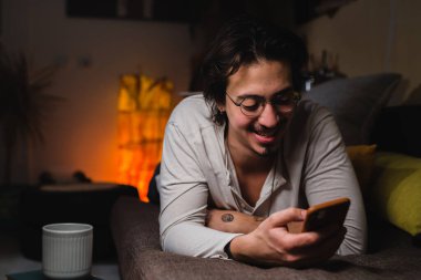 Adam odasındaki kanepede uzanıyor ve cep telefonu kullanıyor.