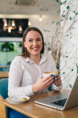 Kafeteryada dizüstü bilgisayarda çalışan ve kahve içen bir kadın.