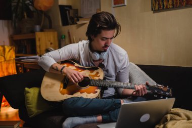 Genç adam odasında akustik gitar çalıyor.