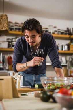 Genç beyaz adam mutfakta salata hazırlarken elinde bir bardak kırmızı şarap tutuyor.