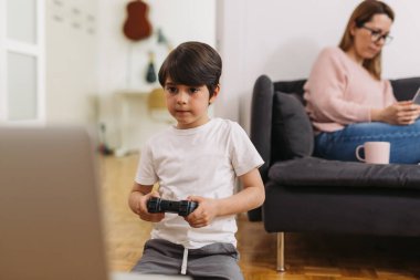 Genç beyaz çocuk elinde joystick ile evde bilgisayar oyunu oynuyor. Annesi arka planda bir kanepede oturuyor.