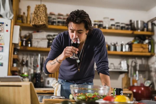 Genç beyaz adam mutfakta yemek hazırlarken kırmızı şarap tadıyor.