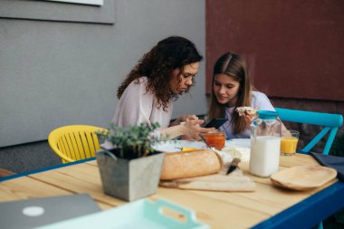 caucasian mother and her young daughter using mobile phone while breakfast outdoor in backyard