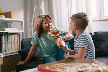 Anne ve oğlu pizza yiyor ve oturma odasında konuşuyorlar.