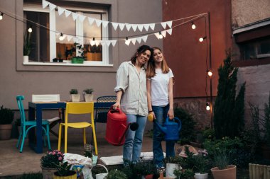 mother and daughter standing in backyard holding watering can and looking at camera. they are gardening plants in backyard