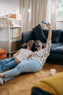 mother and daughter with cosmetic masks applied relaxing at home and taking selfie with smartphone