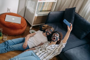mother and daughter with cosmetic masks applied relaxing at home and taking selfie with smartphone