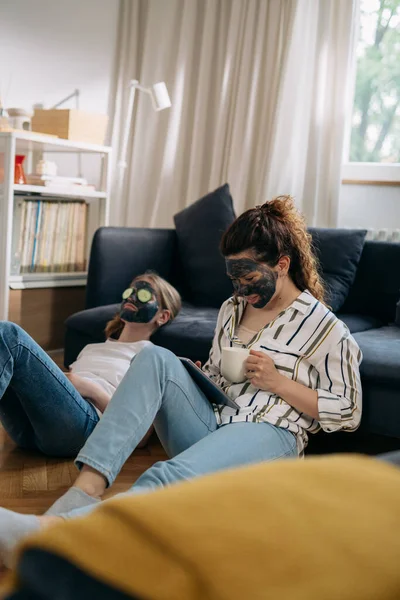 mother and her daughter with cosmetic masks applied on their faces relaxing at home. mother drinking coffee and using digital tablet.