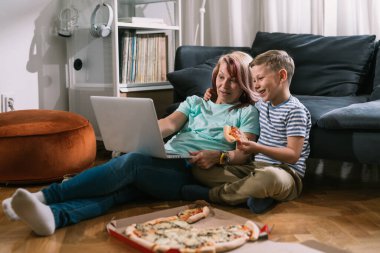 Anne ve oğlu dizüstü bilgisayardan film izliyor ve evde pizza yiyorlar.