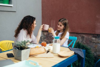 young mother and daughter cheers while having breakfast in backyard on terrace