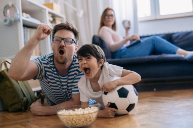 Baba ve oğul yerde yatar ve futbol maçı izler ve evdeki favori takımları için tezahürat yaparlar.