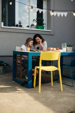 mother and daughter eating breakfast and talking in their home backyard. they are using smartphone