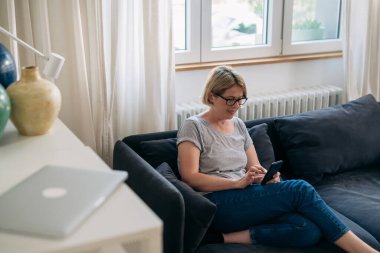 Orta yaşlı bir kadın kanepede oturur ve oturma odasında akıllı telefon kullanır.