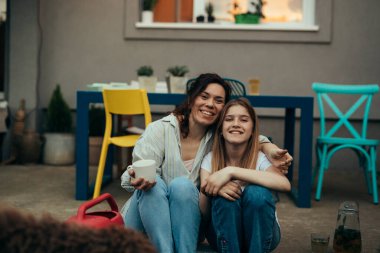 Anne-kız kucaklaşıp kameraya bakarken terasta oturuyorlar.