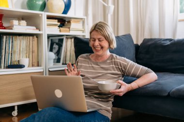 Kadın oturma odasından mısır gevreği kahvaltısı yaparken bilgisayarını video görüşmesi için kullanıyor.