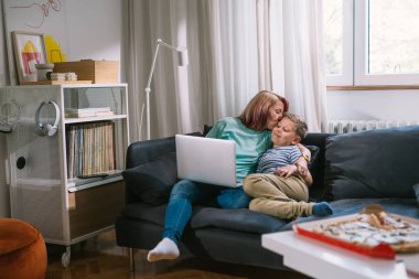 Anne, oturma odasında kanepede otururken oğlunu öpüyor.