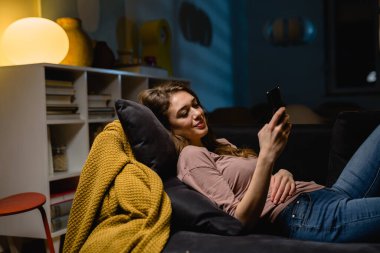 Kanepede dinlenen ve akıllı telefondan mesaj atan bir kadın. Akşam sahnesi