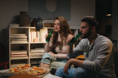 Beyaz bir çift pizza yiyor ve evde bira içiyor.