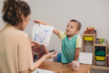 Anaokulunda öğretmenleri resim yapan çocuklar. Çocuk işini gösteriyor.
