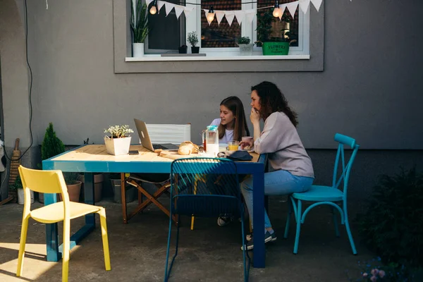 Anne ve kızı evlerinin arka bahçesinde kahvaltı ediyorlar. Yemek yerken dizüstü bilgisayar kullanıyorlar.