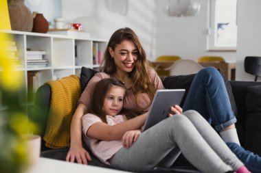 Anne ve kızı oturma odasında kanepede yatıyorlar ve dijital tablet kullanıyorlar.