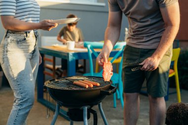 Adam et ve sosisleri barbeküye koyuyor.