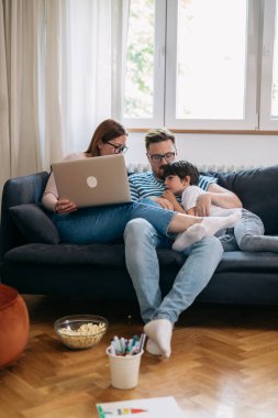 Aile gülümsüyor ve evde dizüstü bilgisayar kullanıyor. Oturma odasındaki kanepede oturuyor.