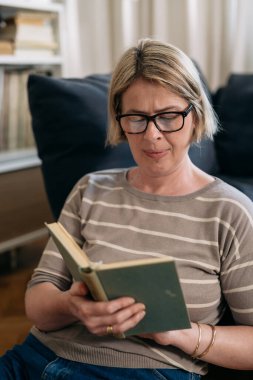 Evde kitap okuyan orta yaşlı bir kadın.