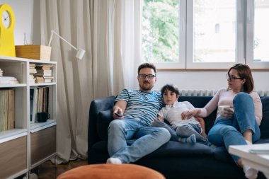 Üç kişilik bir aile kanepede oturuyor ve evde birlikte televizyon izliyorlar.