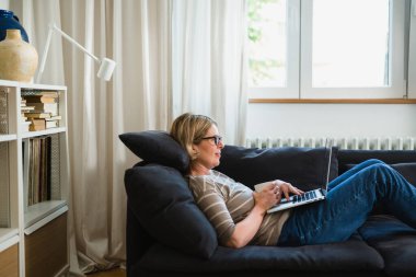 Kadın evinde kanepede uzanıyor ve dizüstü bilgisayar kullanıyor.