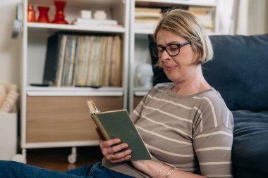 Evde olgun bir kadın kitap okuyor.