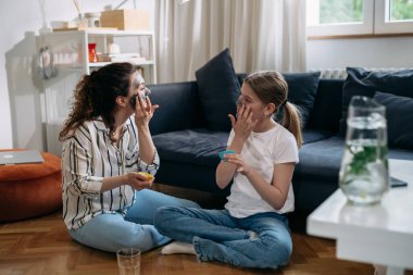 mother and daughter applying cosmetic face mask while sitting in their living room