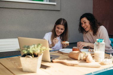 mother and daughter having breakfast together in backyard at home. looking at laptop and laughing