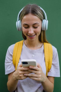 portrait of young female student with headphones and yellow backpack on green background typing smartphone
