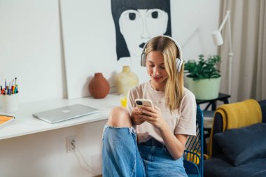 student sits in her room using smartphone and has headphones