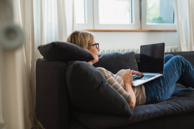 Kadın kanepeye uzanmış ve oturma odasında dizüstü bilgisayar kullanıyor.