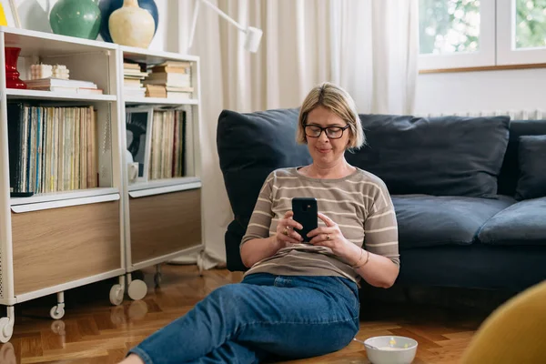 caucasian woman sits on floor in her living room and using mobile phone