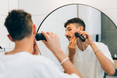 Genç bir adam banyoda elektrikli tıraş makinesi kullanıyor.