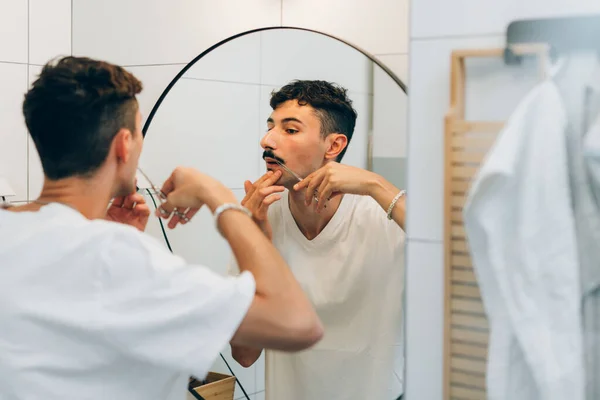 young man shapes his mustaches using scissors in bathroom