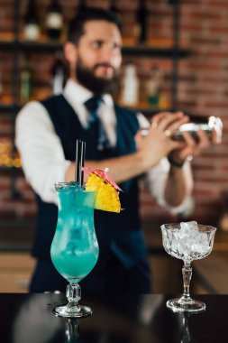 Bar tezgahında mavi lagün kokteyli, bulanık arka planda barmen kokteyli sallıyor.