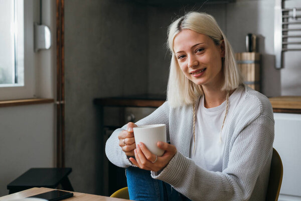 young adult generation z woman sits in her kitchen, drinking coffee and looking at camera