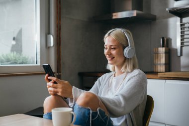 Kadın kulaklıkla müzik dinliyor ve evde akıllı telefon kullanıyor.