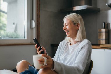 Gülümseyen kadın kahve içiyor ve evinde cep telefonu kullanıyor.