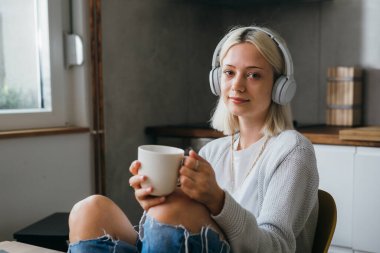 Kulaklıklı genç bir kadın sabah kahvesini yudumlarken kameraya bakıyor.