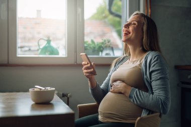 Akıllı telefon kullanan ve evde gülen hamile bir kadın.