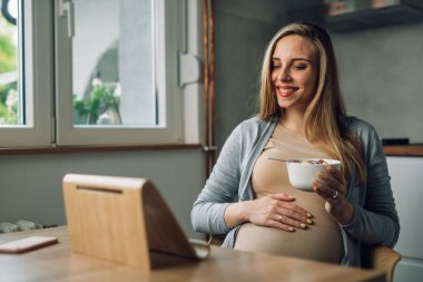 Hamile kadın evde sağlıklı meyve salatası yerken dijital tablet kullanıyor.