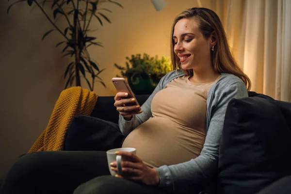 高加索孕妇坐在沙发上 在客厅里用智能手机 — 图库照片